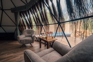Un interior acogedor con piso de madera, un sillón gris, una mesa de centro de madera con un florero, y vistas a una piscina y zona exterior a través de amplios ventanales.