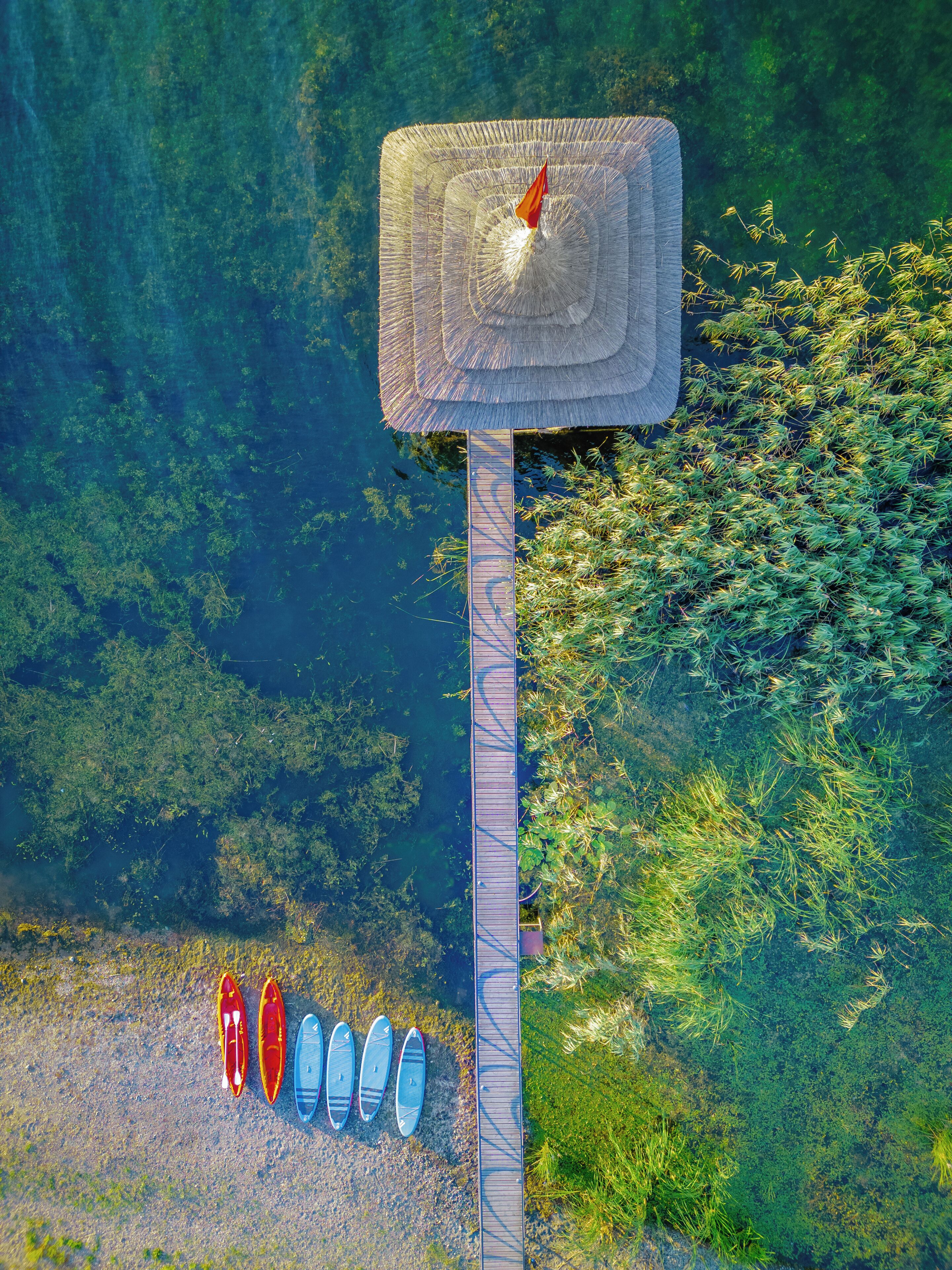 An aerial view of a dock with a thatched roof structure, surrounded by greenery and clear water.