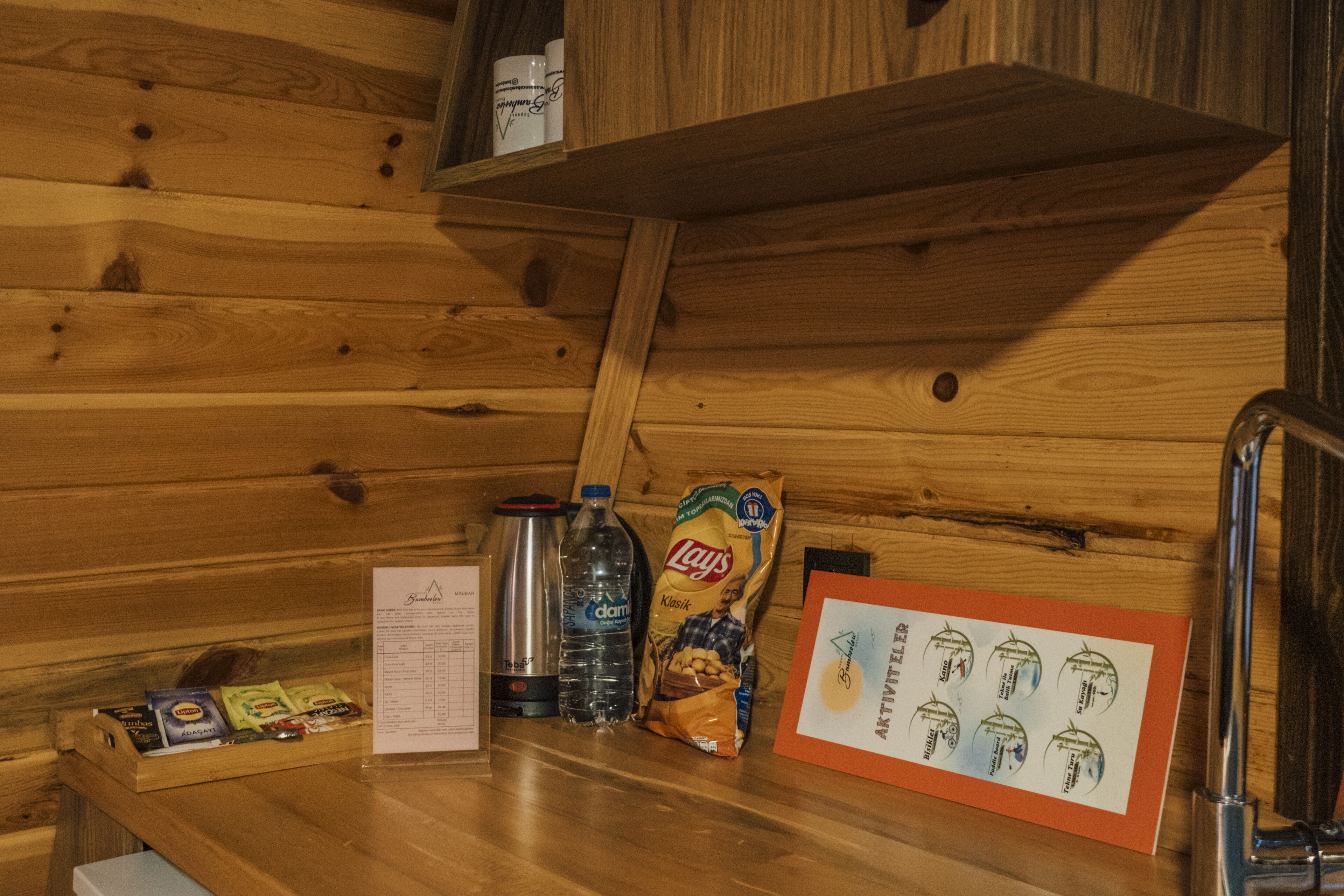 Interior de una cabaña de madera con repisa de madera, una botella de agua de metal, una bolsa de papas Lay's, un termo y un menú.