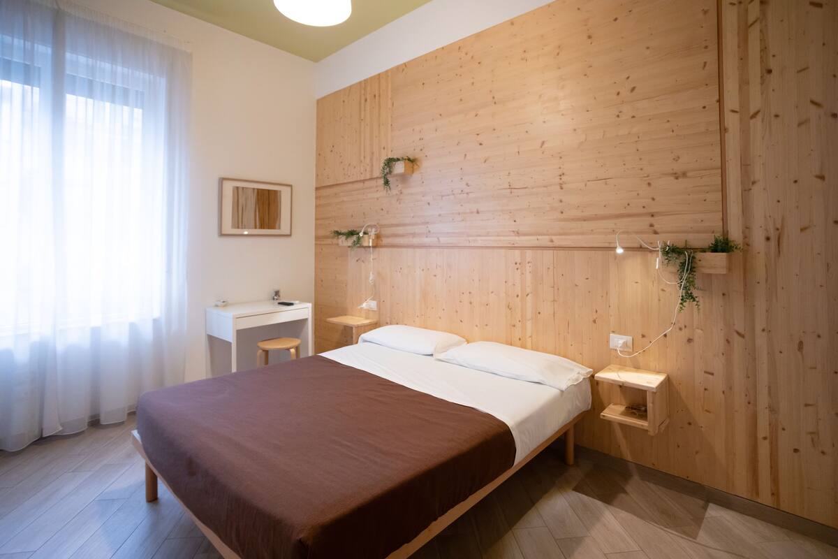A bedroom with a wooden headboard, a bed with white linens, a bedside table, a small desk, and a window with sheer curtains.