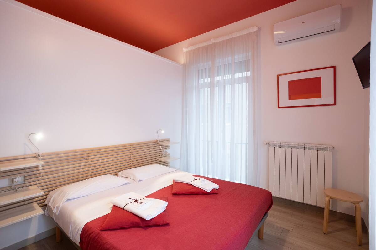A modern hotel room with a large bed, a bedside table, a chair, a window with sheer curtains, and a radiator.