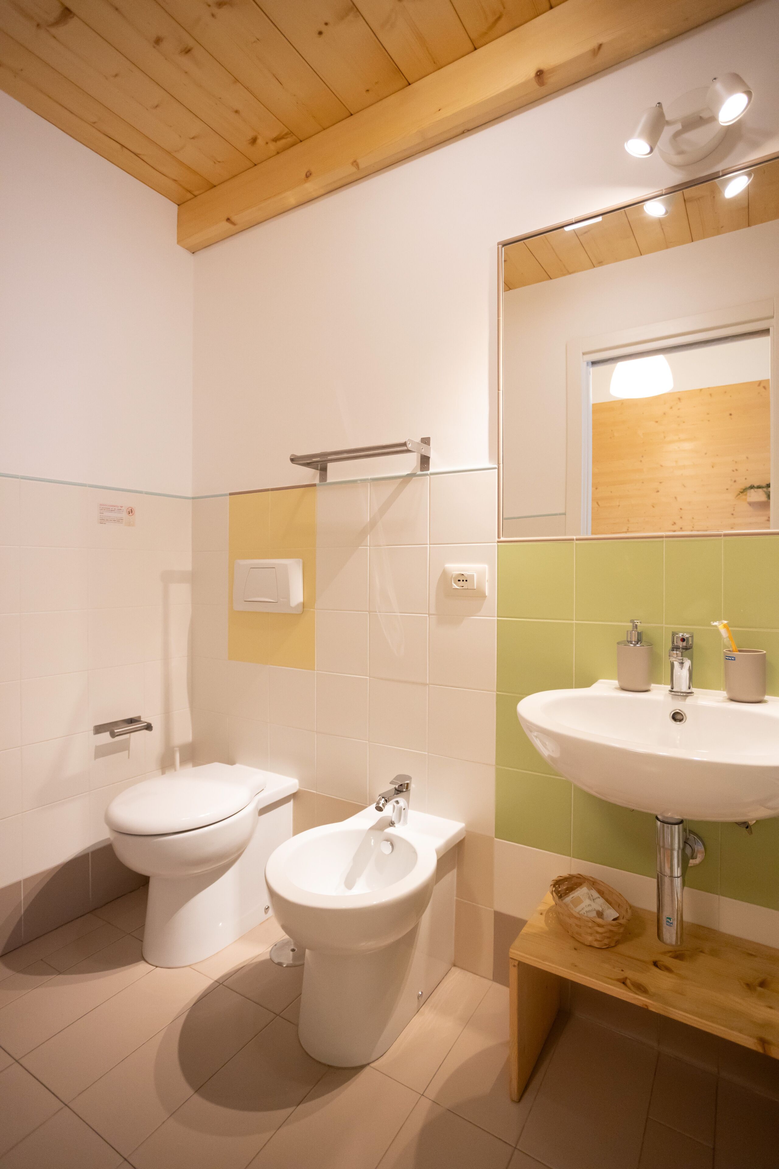 A bathroom with a toilet, bidet, and sink, wooden ceiling, and a mirror with a wooden frame.