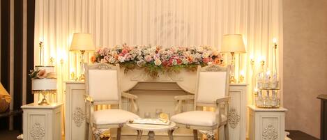 A room with two white upholstered chairs, a small round table, and a decorative floral arrangement above a fireplace.