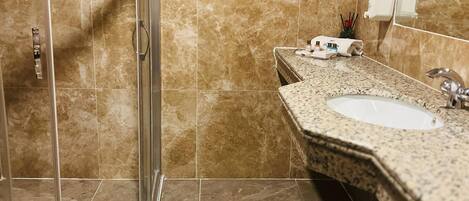 A bathroom with a glass shower enclosure, a sink with a marble countertop, and a hand soap dispenser on the wall.