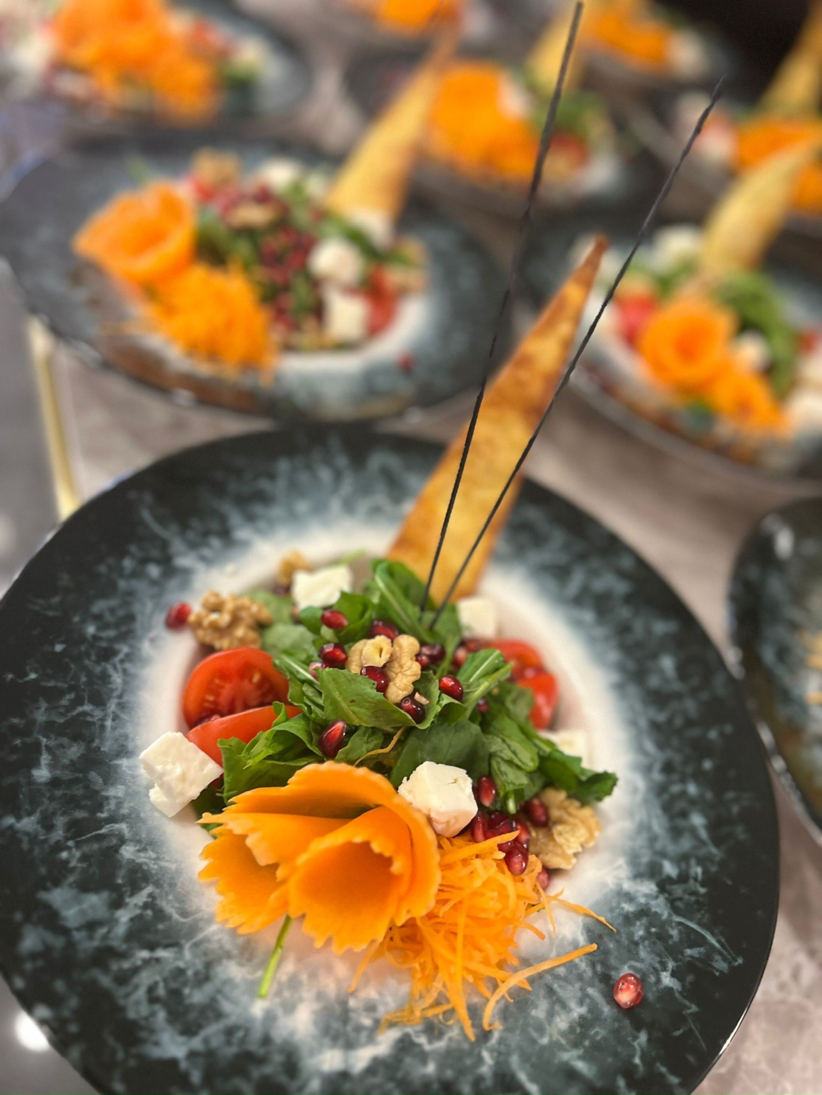 A close-up of a gourmet dish with a variety of ingredients, including greens, nuts, and a decorative orange element, served on a dark plate.