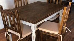 Ein Esstisch mit sechs Holzstühlen, jeweils mit weißem Kissen. Der Tisch hat eine dunkle Holzplatte und weiß gestrichene Beine. Der Raum verfügt über Holzboden und eine weiße Ziegelwand.