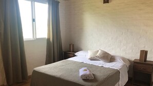 Ein ordentlich bezogenes Bett mit weißer Decke und Kissen, ein zusammengerolltes Handtuch auf dem Bett, ein Nachttisch aus Holz mit einer Lampe und ein Fenster mit Vorhängen.