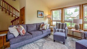 A living room with a sofa, armchair, and ottoman, a staircase, and large windows with a view of the outdoors. - YANK01 - Cozy Home, Gas Fireplace, Dog Friendly (Sunriver)