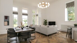 Een moderne eethoek met een houten visgraatvloer, een centrale eettafel met stoelen, een grijze bank en een televisie aan de muur.