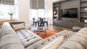 A modern living room with a large grey sofa, a dining table with chairs, and a flat-screen TV mounted on the wall. - Rowley House at Stone Cross Mansion (Ulverston)