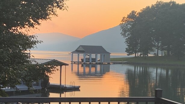 A wooden deck with a view of a calm lake, a dock, and a small building in the distance.