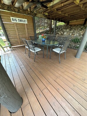 Une terrasse extérieure couverte, avec une table et des chaises, une pancarte sur laquelle on peut lire « GRAND SOURIRE ! » et un mur de pierre en arrière-plan.