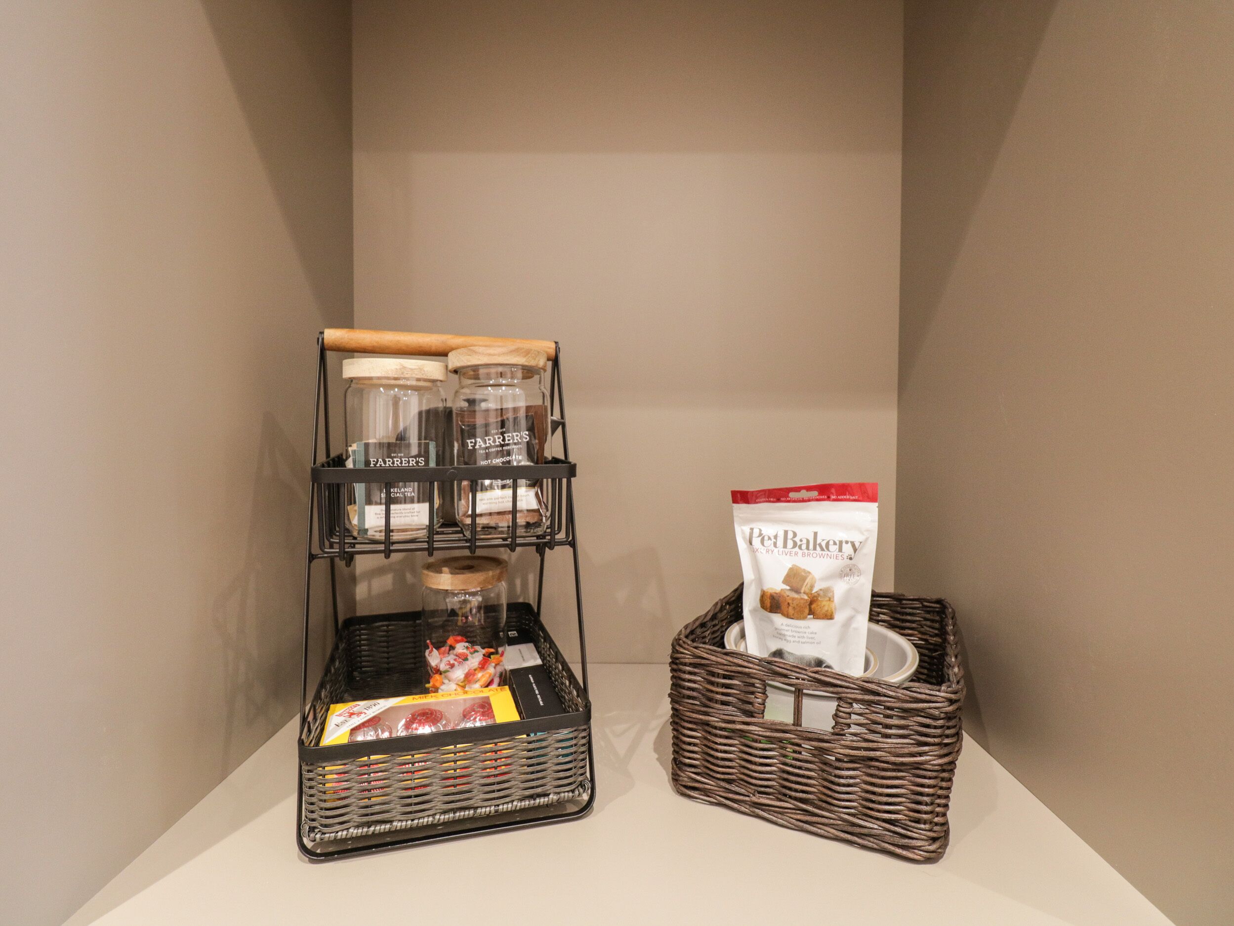 A kitchen area with a black metal rack holding jars and a wicker basket containing a bag of Pet Bakery liver brownies.