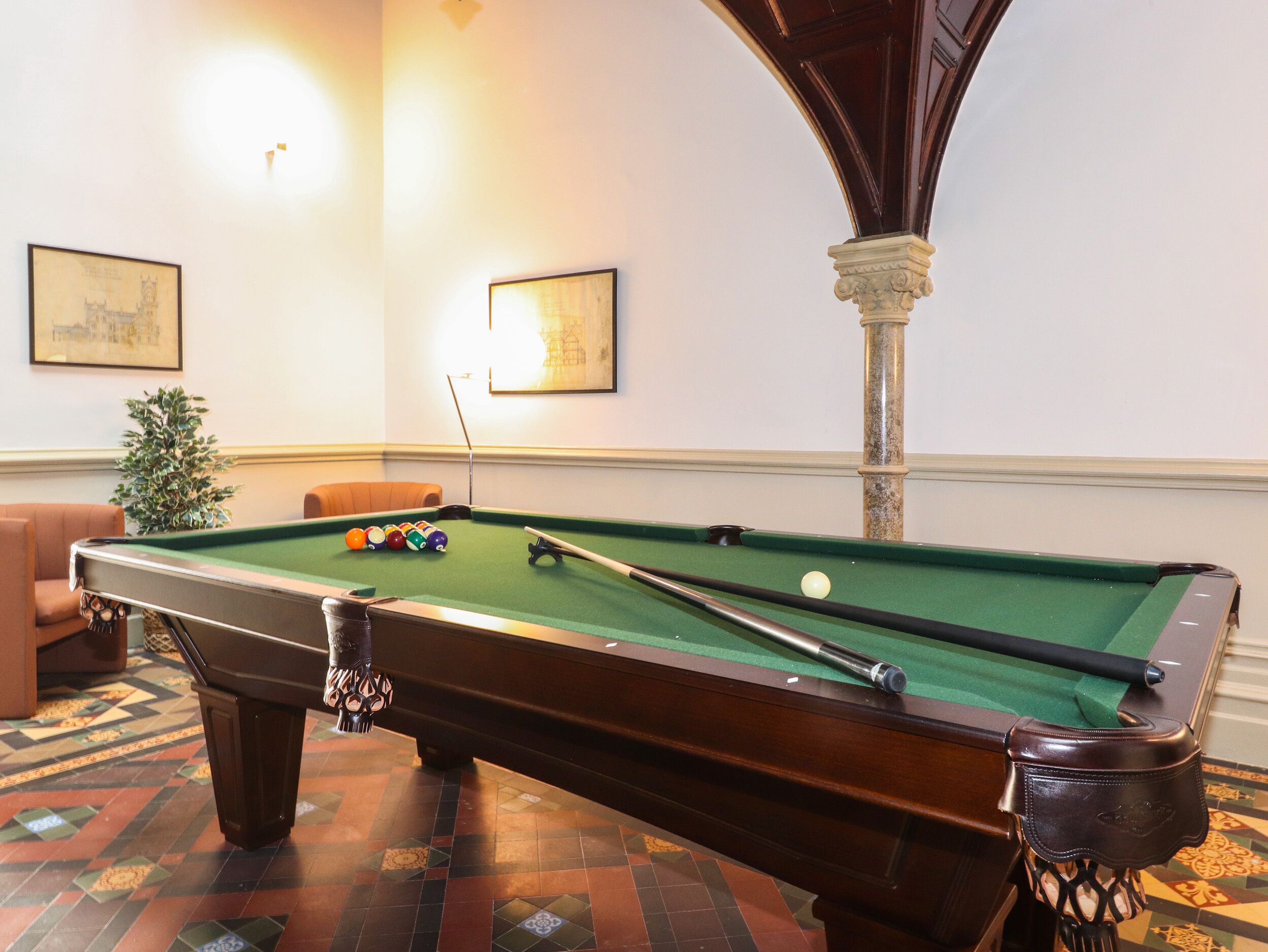 A pool table in a room with patterned flooring, a potted plant, and framed artwork on the wall.