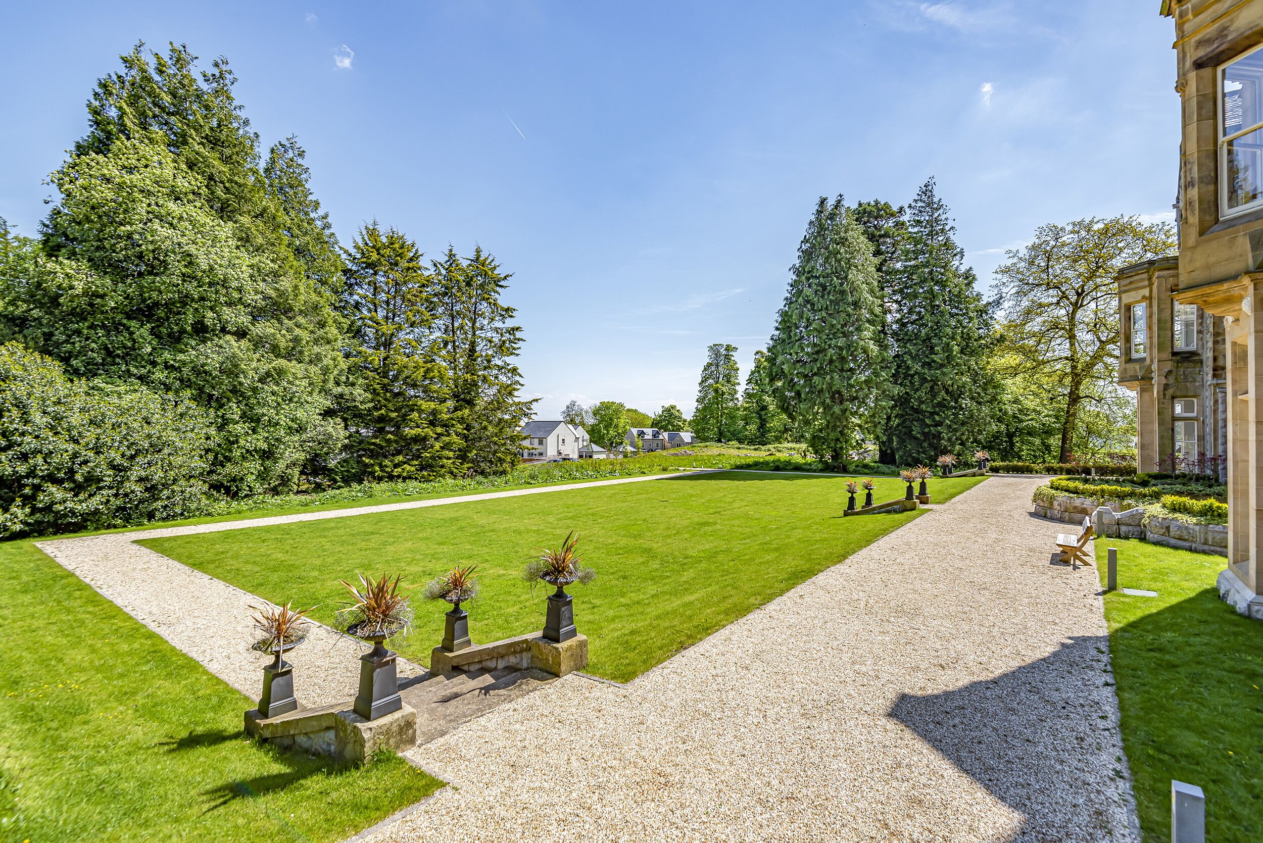 A well-maintained garden with a gravel path leading to a lawn area, surrounded by trees and a building with large windows.