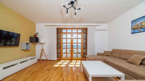 A living room with a wooden floor, a white coffee table, a brown sofa, a TV mounted on the wall, and a large window with curtains. - Holiday apartment with 4 bedrooms and a balcony (Crikvenica)