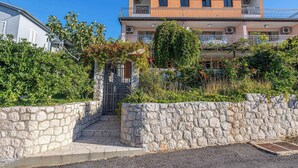 A two-story residential building with a stone wall entrance, a balcony on the upper floor, and lush greenery around. - Holiday apartment with 4 bedrooms and a balcony (Crikvenica)