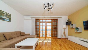 A living room with a beige sofa, a white coffee table, wooden flooring, a large window with blinds, and a ceiling light fixture. - Holiday apartment with terrace and air conditioning (Crikvenica)