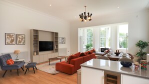 A modern living room with a flat-screen TV, a sofa, a dining area with a table set for two, and a kitchen area with a built-in oven. - The Kathleen Suite Stone Cross Mansion (Ulverston)
