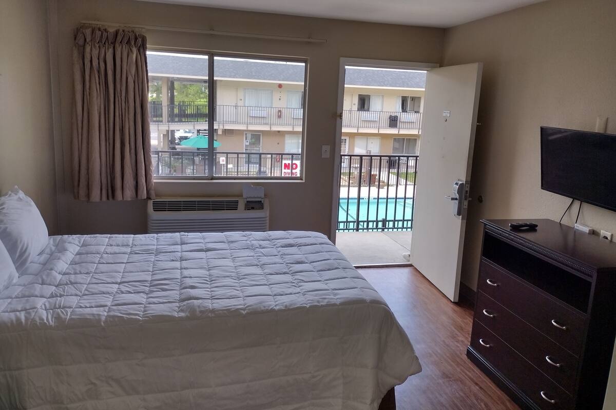 A hotel room with a bed, a TV, a dresser, and a view of a swimming pool and buildings.