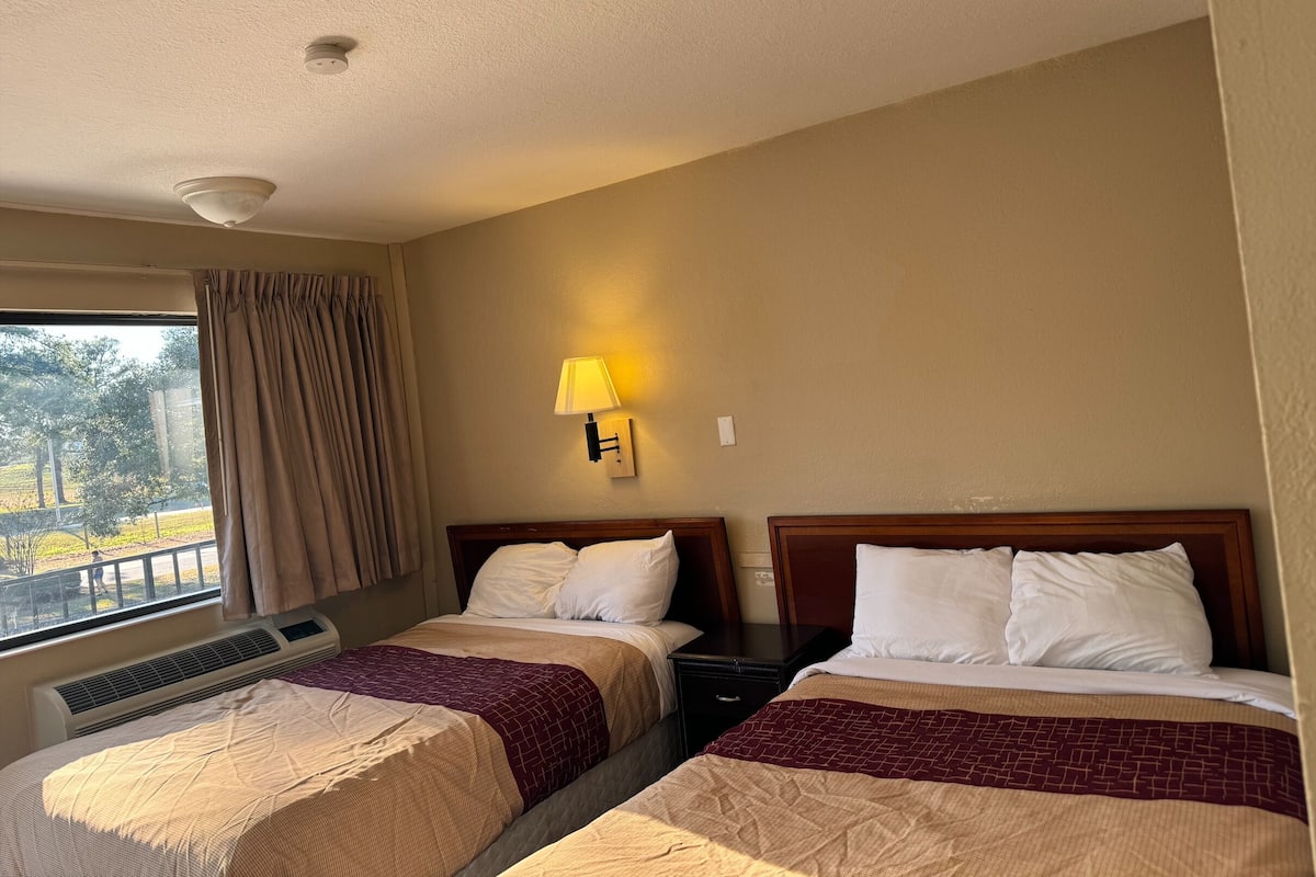A hotel room with two beds, a window with curtains, a wall-mounted lamp, and an air conditioning unit.