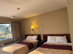A hotel room with two beds, a window with curtains, a wall-mounted lamp, and an air conditioning unit.