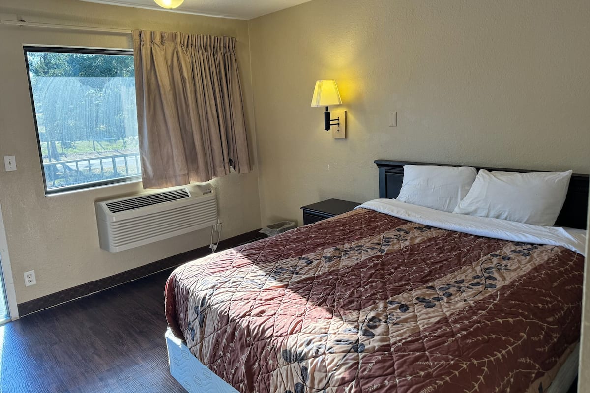 A hotel room with a bed, a window with curtains, an air conditioning unit, a nightstand, and a wall-mounted lamp.