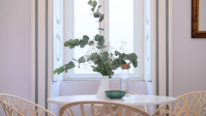 Una mesa redonda con un florero blanco y un cuenco verde, rodeada por cuatro sillas de diseño elaborado, ubicada frente a una ventana con cortinas traslúcidas.