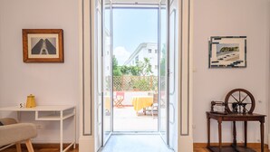 Una habitación con piso de madera, un escritorio blanco, una silla, una mesita con un carrete de hilo y una puerta que da a un espacio al aire libre con una mesa y sillas.