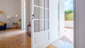 Una habitación con suelo de madera, una puerta blanca con paneles de vidrio, un sofá azul, una mesita con una lámpara, y vista a un jardín a través de un arco.