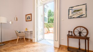 Una habitación con piso de madera, un escritorio con silla, una lámpara, un cuadro enmarcado, una mesita con ruedas y una puerta que da a un espacio al aire libre.