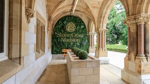 Um corredor arqueado com colunas de pedra e um banco, conduzindo a uma placa com a inscrição 'Stone Cross Mansion'.