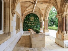 Un corredor arqueado con columnas de piedra y un banco, que conduce a un letrero que dice 'Stone Cross Mansion'.