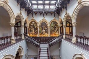 Una majestuosa escalera con barandillas ornamentadas y un tragaluz arriba, que conduce a una galería con pinturas clásicas.