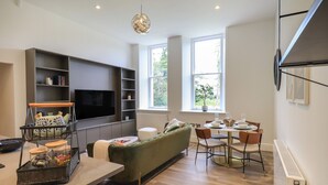 Uma sala de estar moderna com televisão de tela plana, um sofá verde, área de jantar com mesa posta para quatro pessoas e uma janela grande que oferece iluminação natural.