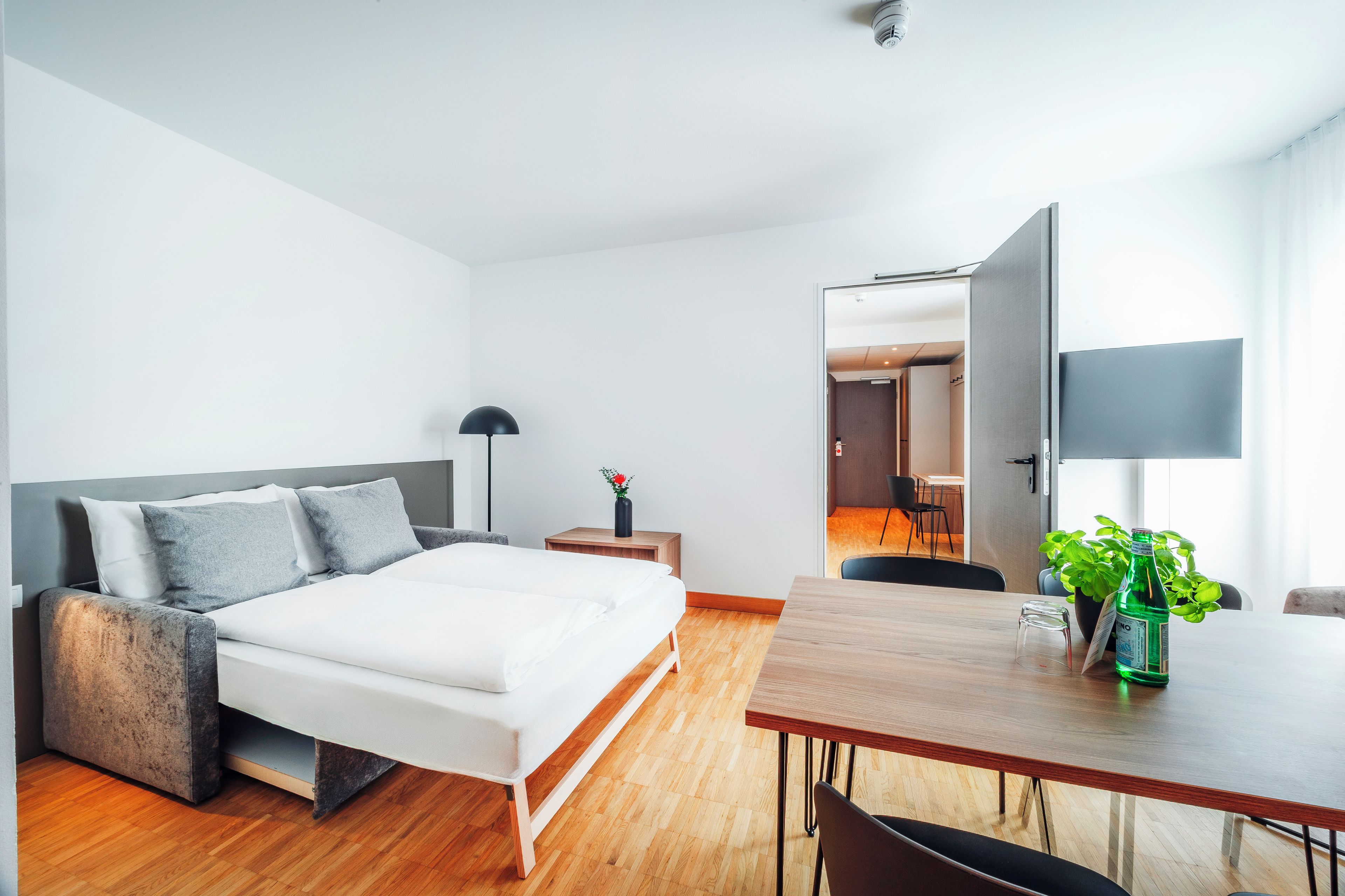 Una moderna camera d'albergo con un letto, un comodino, una lampada e un'area ristoro con tavolo e sedie.