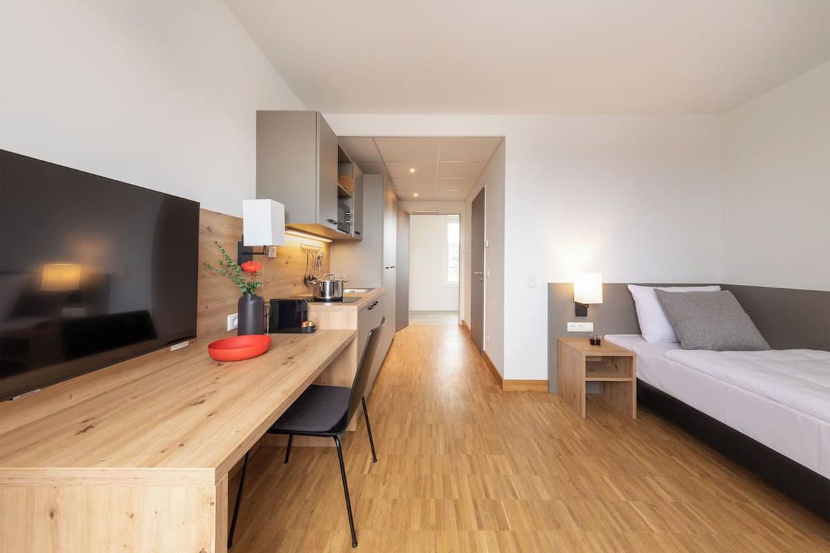 A modern hotel room with a wooden desk, a bed, a TV, and a kitchenette.