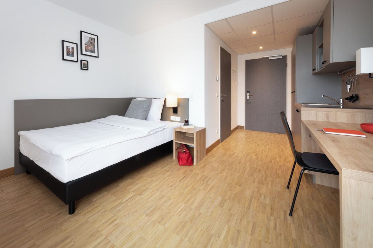 A modern hotel room with a bed, bedside table, lamp, and a small red bag. There is a wooden desk with a chair and a kitchen area in the background.