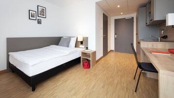 A modern hotel room with a bed, bedside table, lamp, and a small red bag. There is a wooden desk with a chair and a kitchen area in the background.