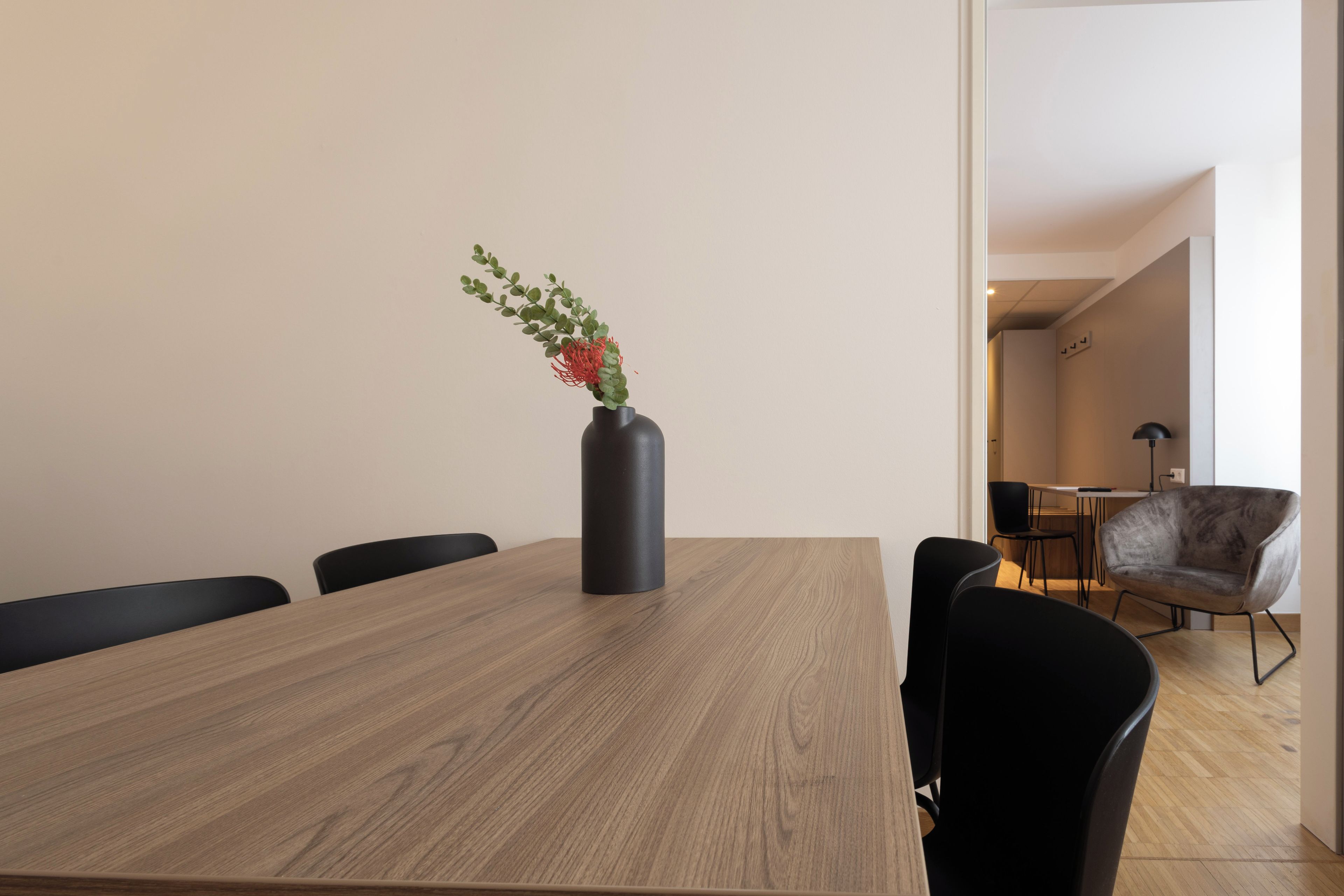 Un salón de conferencias con una mesa de madera, sillas negras y un jarrón con follaje verde y flores rojas.