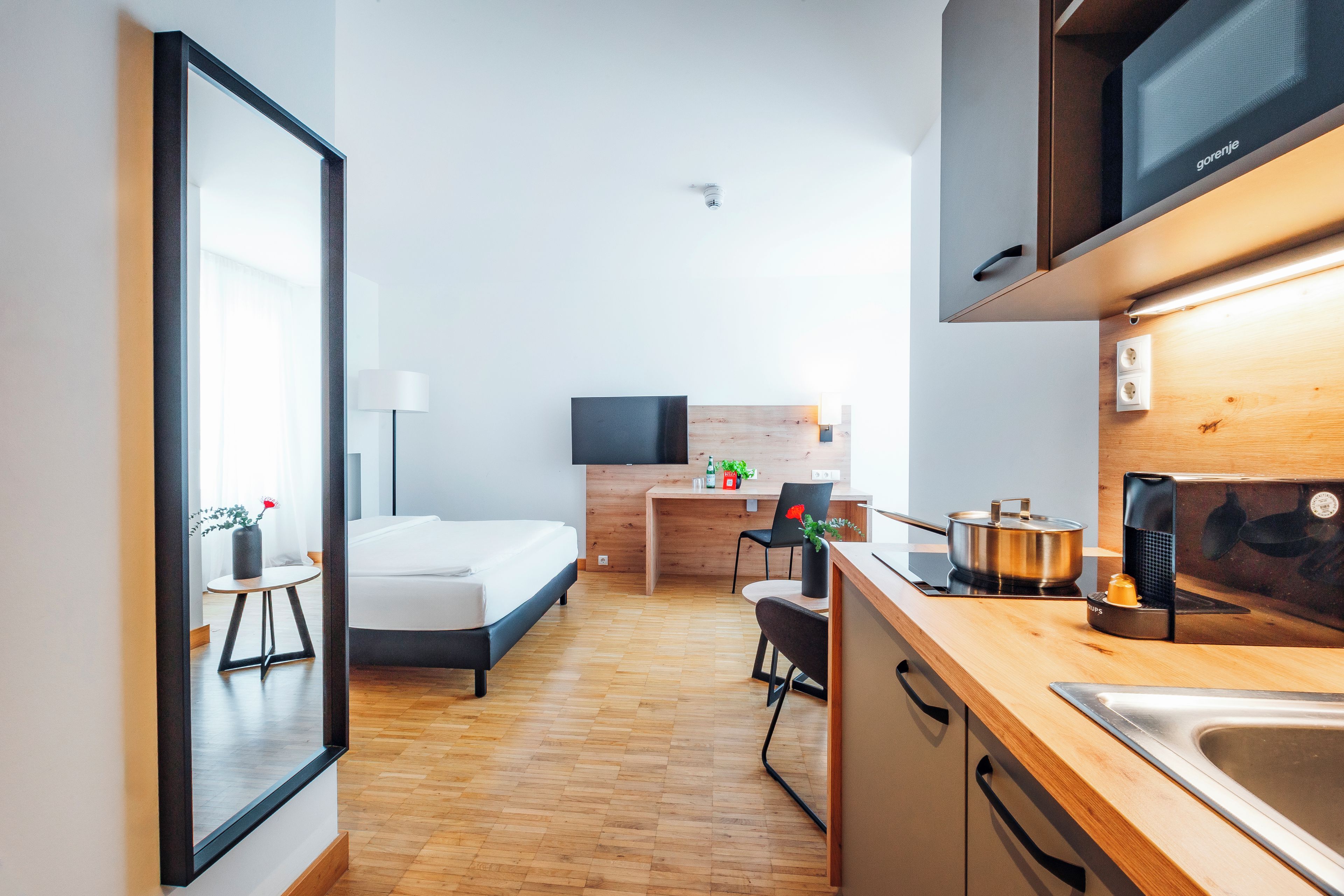 Habitación de hotel moderna con cama, escritorio, silla, televisor y cocina equipada con estufa y lavaplatos, además de suelo de madera.