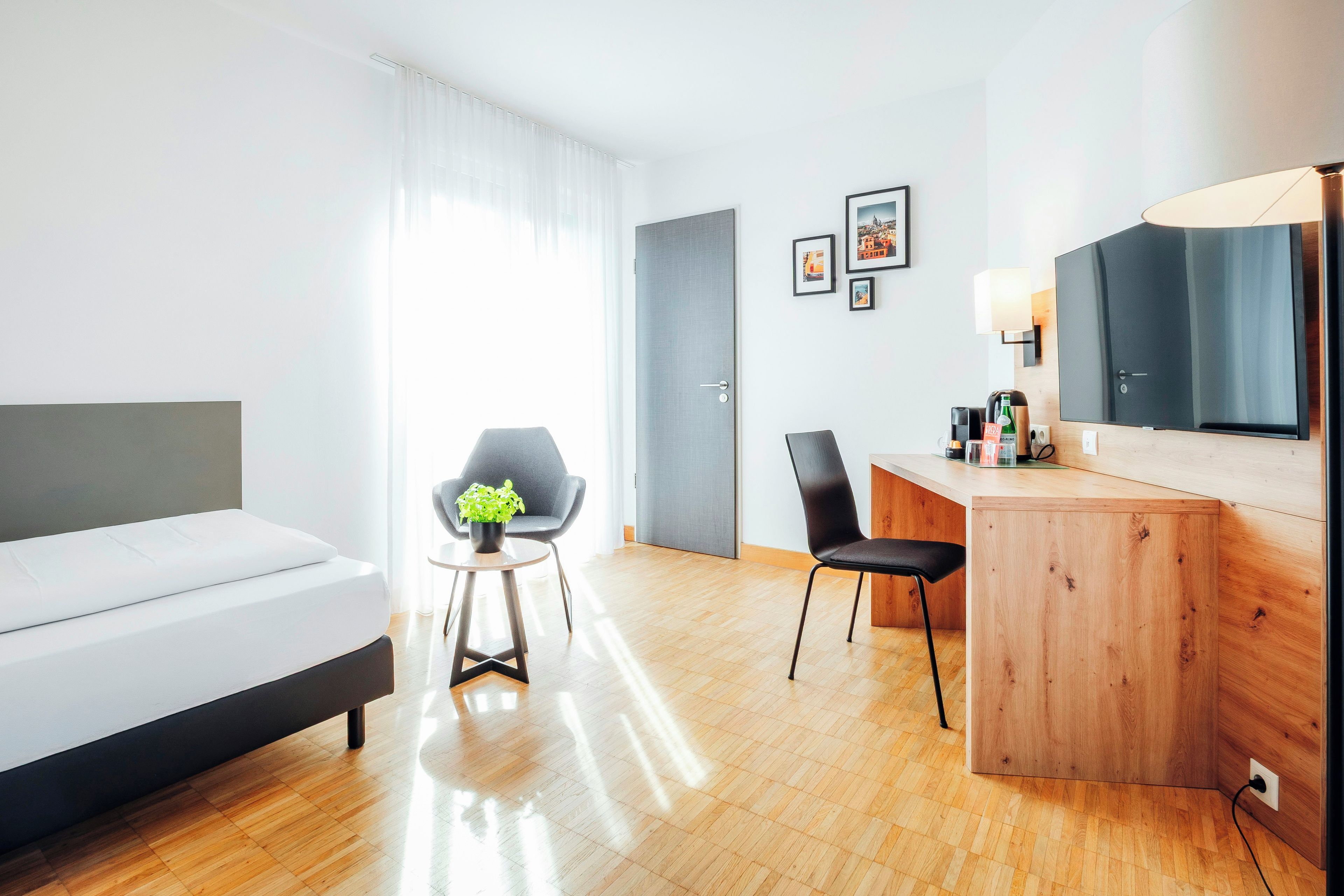 A modern hotel room with a bed, a desk, a chair, a small table with a plant, and a mirror.