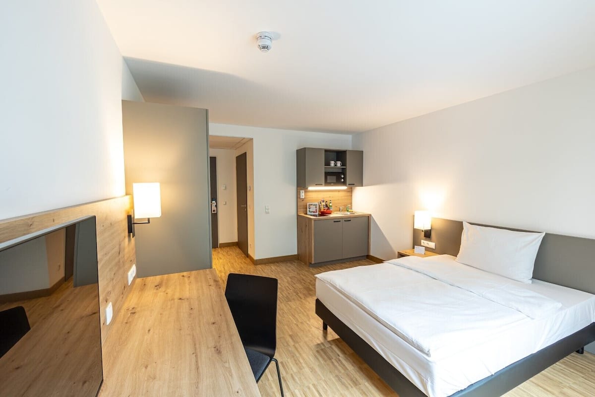 A modern hotel room with a large bed, a desk, and a kitchenette.