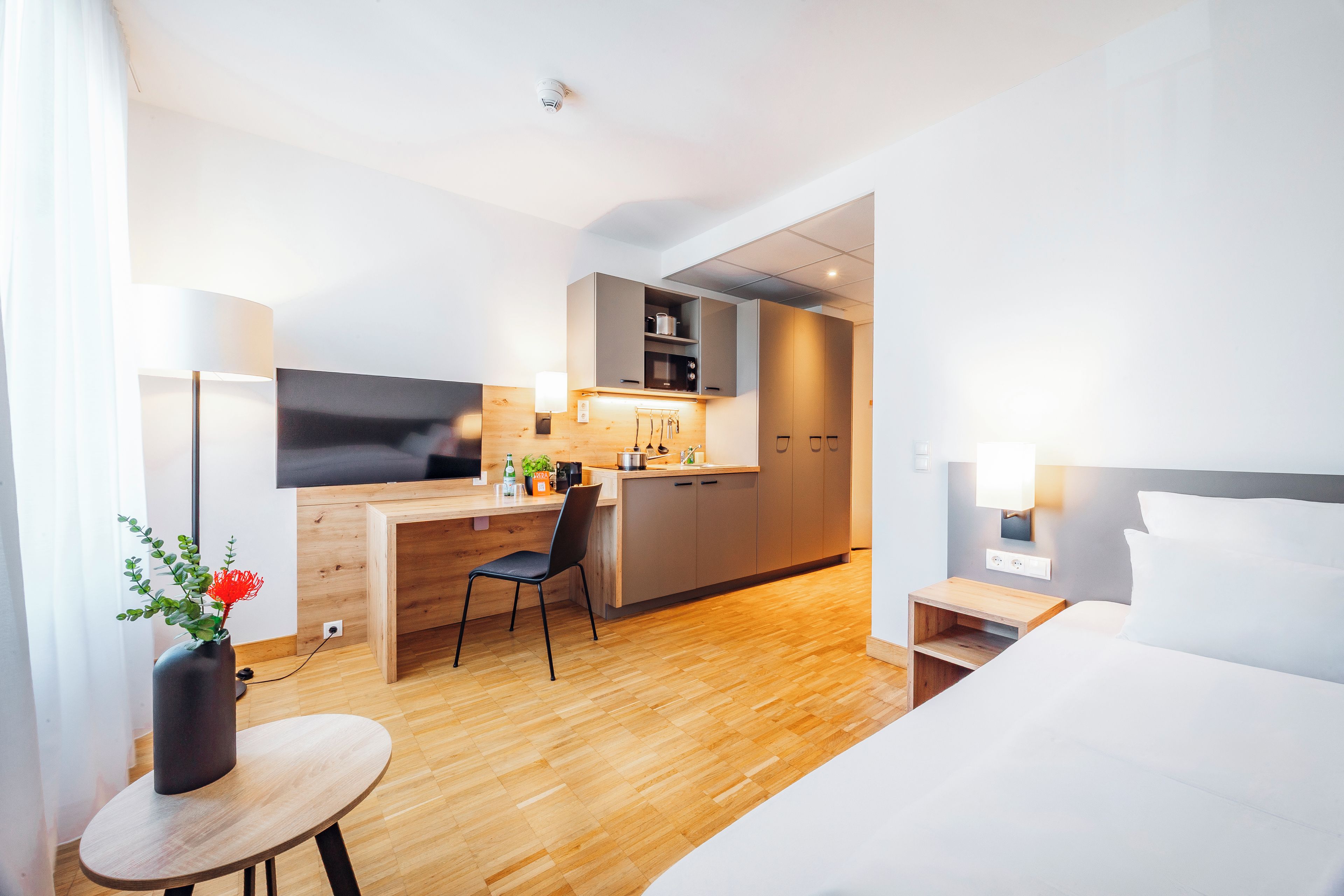 A modern hotel room with a bed, a desk, a chair, a small table, and a kitchenette.