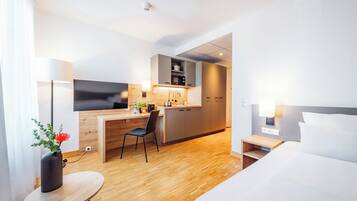 A modern hotel room with a bed, a desk, a chair, a small table, and a kitchenette.