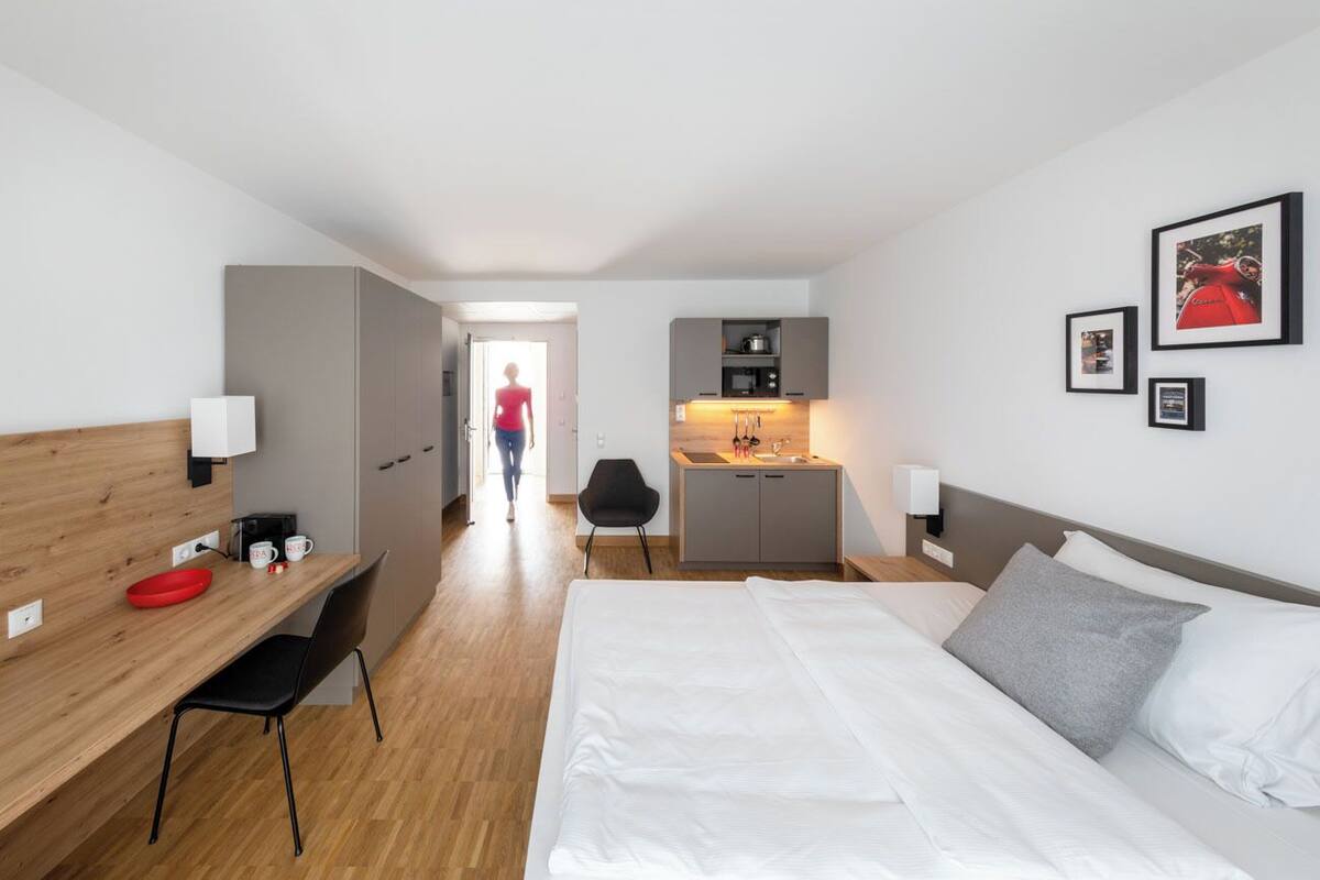 A modern hotel room with a wooden desk, a bed with white linens, a kitchenette, and a view of the hallway.