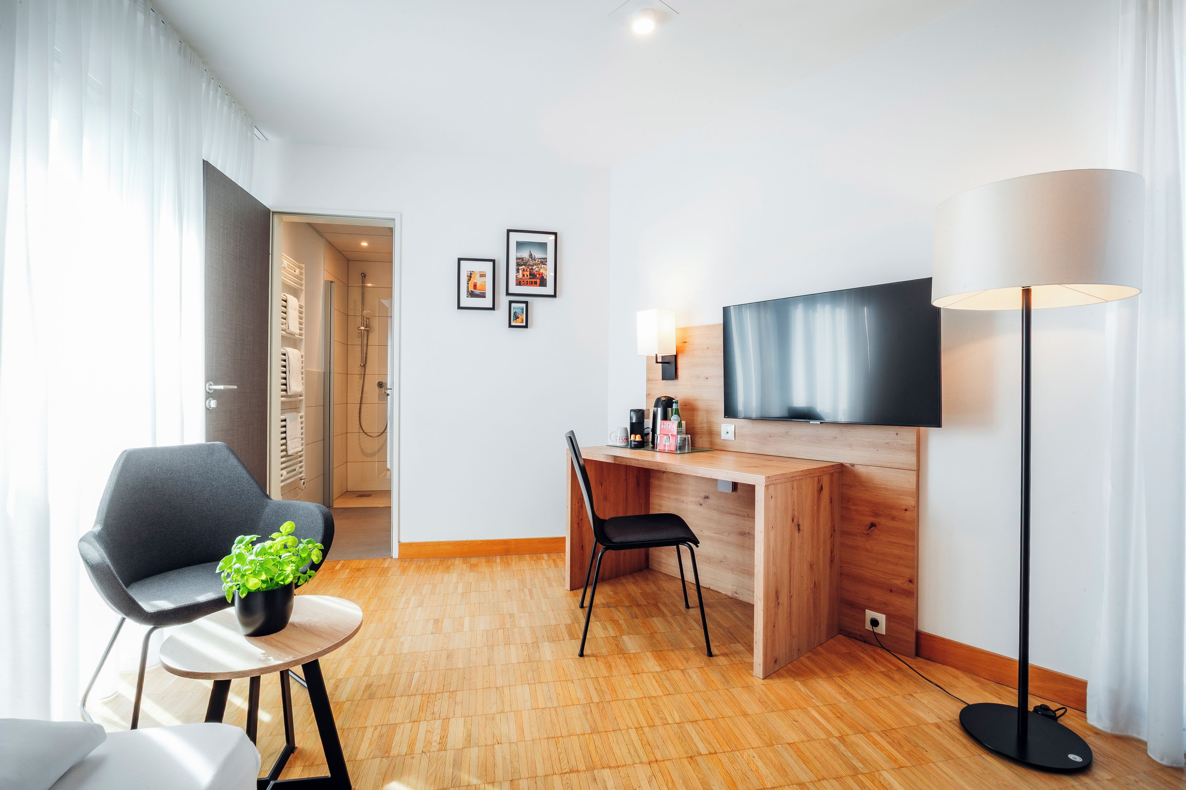 Ein modernes Hotelzimmer mit einem Holzbüro, einem Flachbildfernseher, einer Stehlampe, einem Sessel und einem kleinen Tisch mit einer Pflanze.