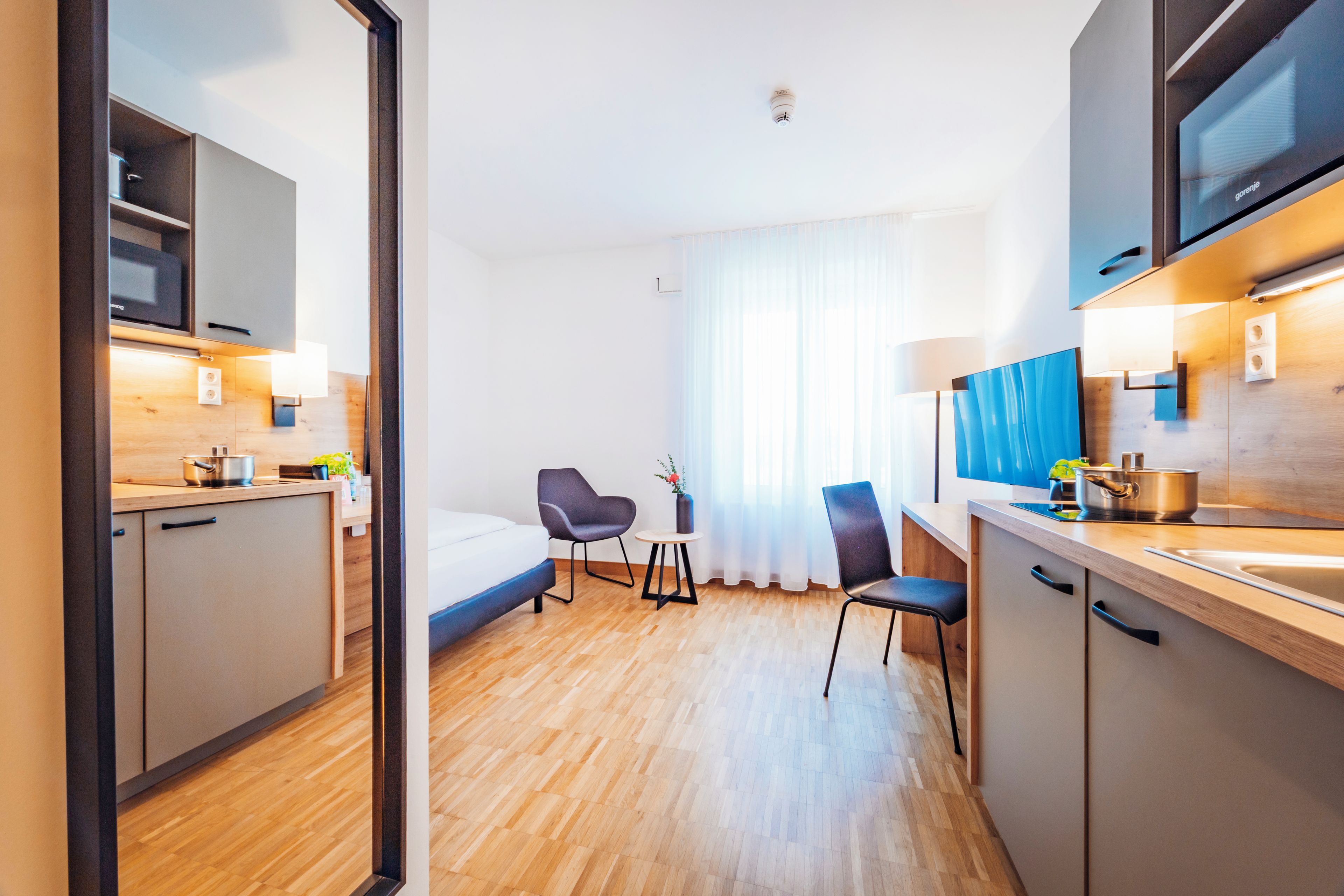 A modern hotel room with a kitchenette, a bed, a chair, and a large window with curtains.
