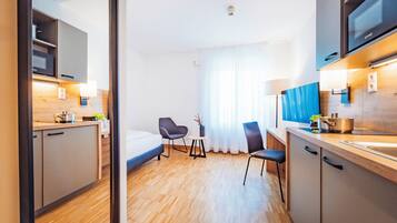 A modern hotel room with a kitchenette, a bed, a chair, and a large window with curtains.