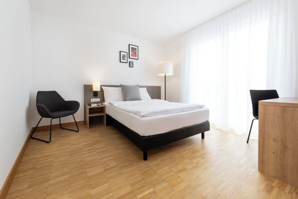 A modern hotel room with a bed, two chairs, a desk, and a nightstand with lamps.